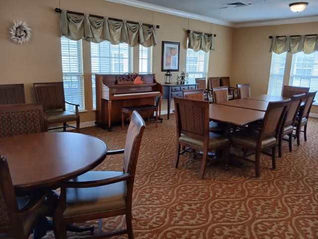 A well-lit room with multiple wooden tables and chairs arranged on a patterned carpet. There is a piano against the wall with a small bench in front of it. The walls have several windows with beige curtains, a framed picture, and decorative items on a side table.