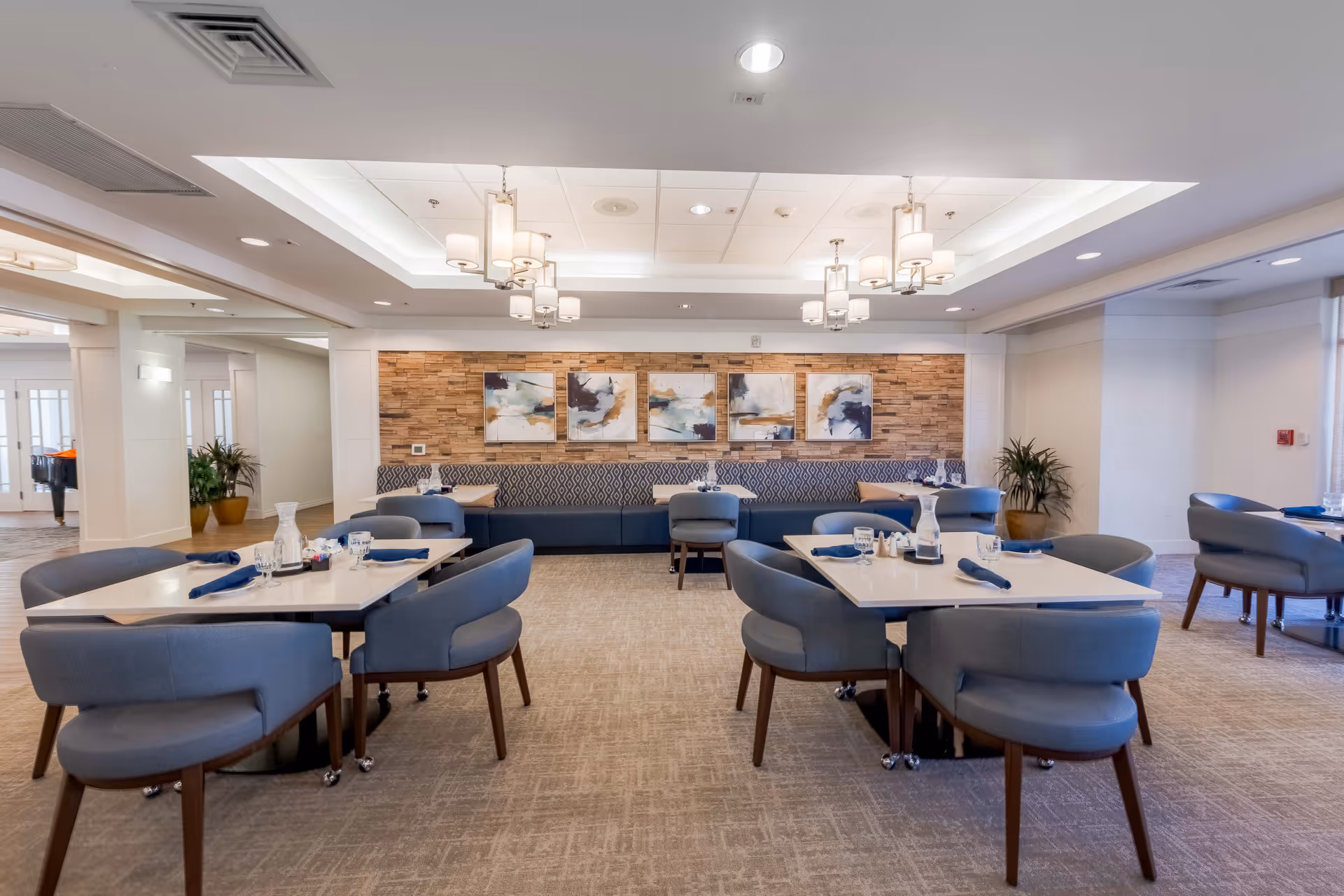 A bright and modern dining room with multiple square tables set with blue napkins, glassware, and condiments. The room features comfortable blue upholstered chairs, a long cushioned bench along a wall with decorative abstract paintings, and contemporary ceiling light fixtures. There are potted plants in the corners and large windows allowing natural light to fill the space.