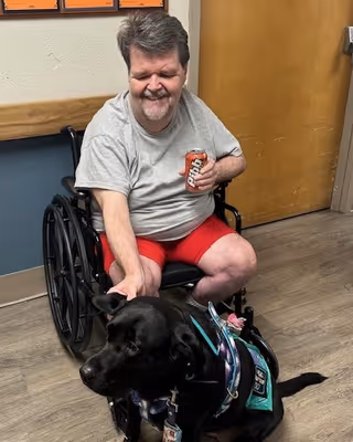 A man sitting in a wheelchair indoors, smiling and holding a can of soda in one hand while petting a black service dog wearing a vest.