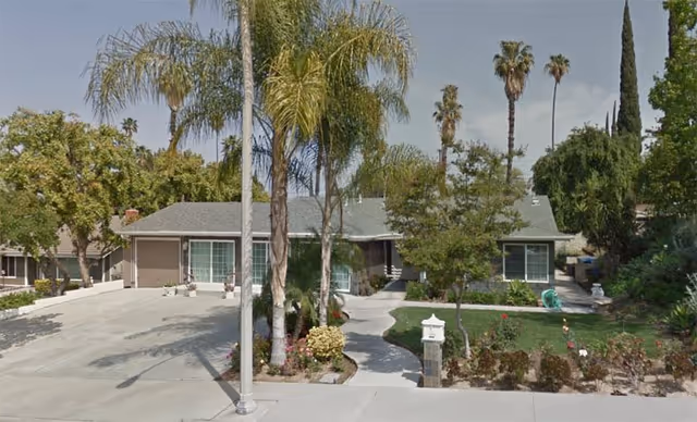 Front exterior view of a single-story residential building with a driveway, palm trees, and a landscaped garden area with bushes and flowers.