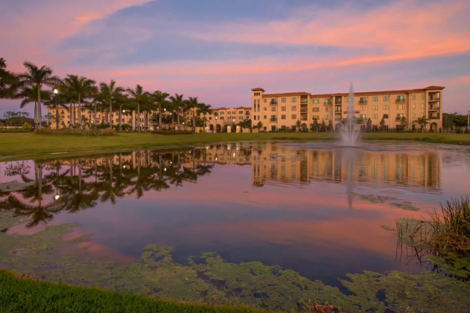 A large multi-story residential building with balconies is reflected in a pond with a central water fountain. The scene is set during sunset with a pink and purple sky. Palm trees line the area between the building and the pond.