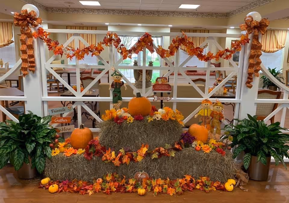 Indoor autumn-themed decoration with hay bales, pumpkins, colorful fall leaves garlands, and scarecrow dolls arranged in front of a white lattice divider. Two potted green plants flank the display, and a dining area with tables and chairs is visible in the background.