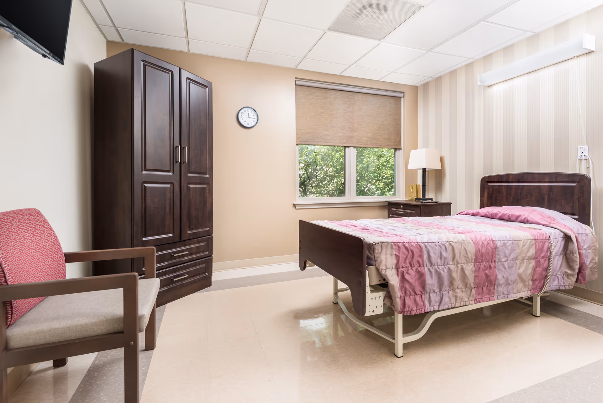 A simple, clean bedroom in a healthcare facility with a single bed covered in a pink and beige quilt, a wooden nightstand with a lamp, a large dark wooden wardrobe, a wall clock, a window with a beige roller shade, and a wooden chair with a red cushion.