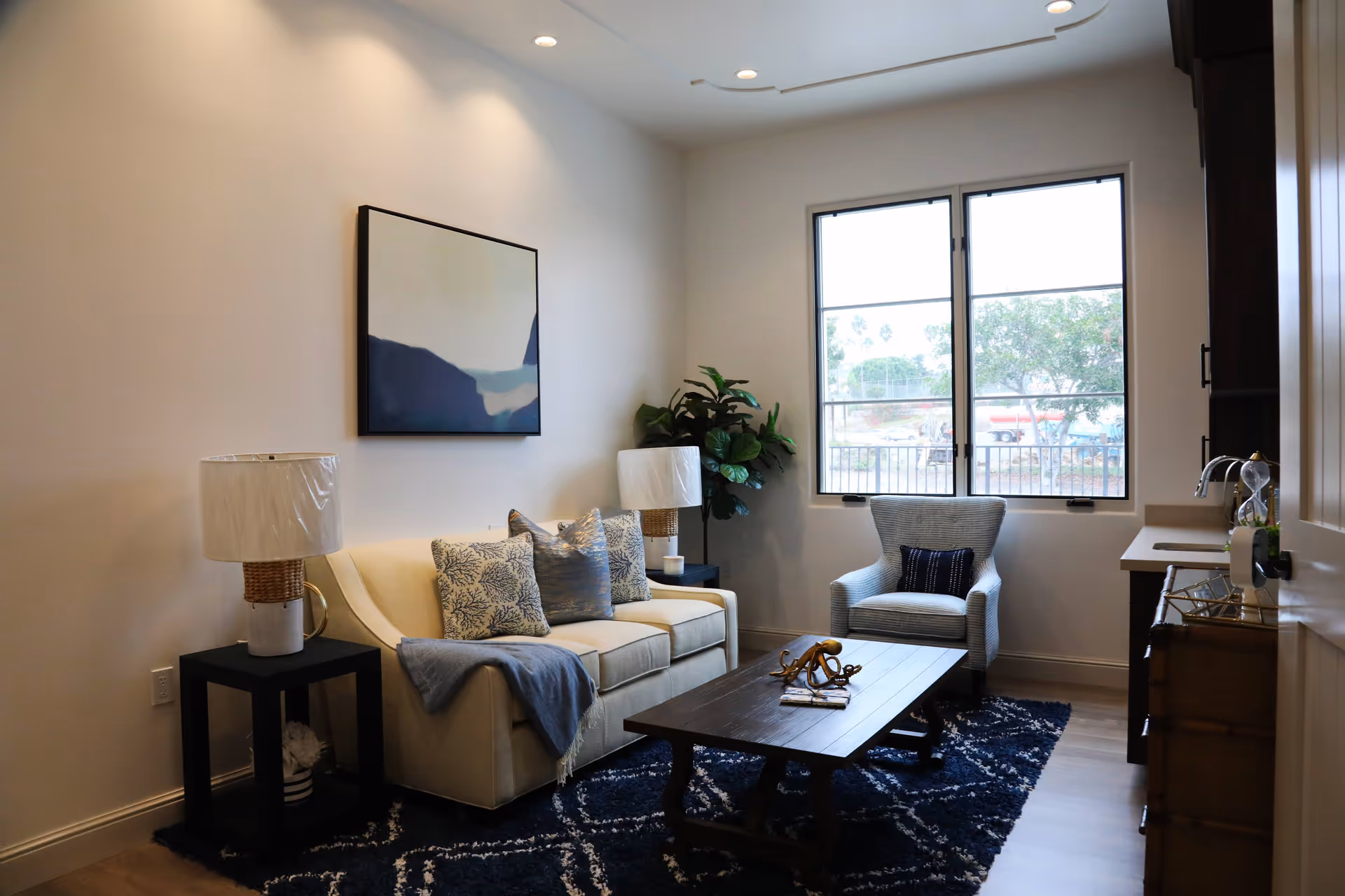 A cozy living room with a beige sofa adorned with decorative pillows and a blue throw blanket, a wooden coffee table with a decorative octopus sculpture, a blue patterned rug, a single armchair, two side tables with lamps, a large window letting in natural light, and a piece of abstract art on the wall.