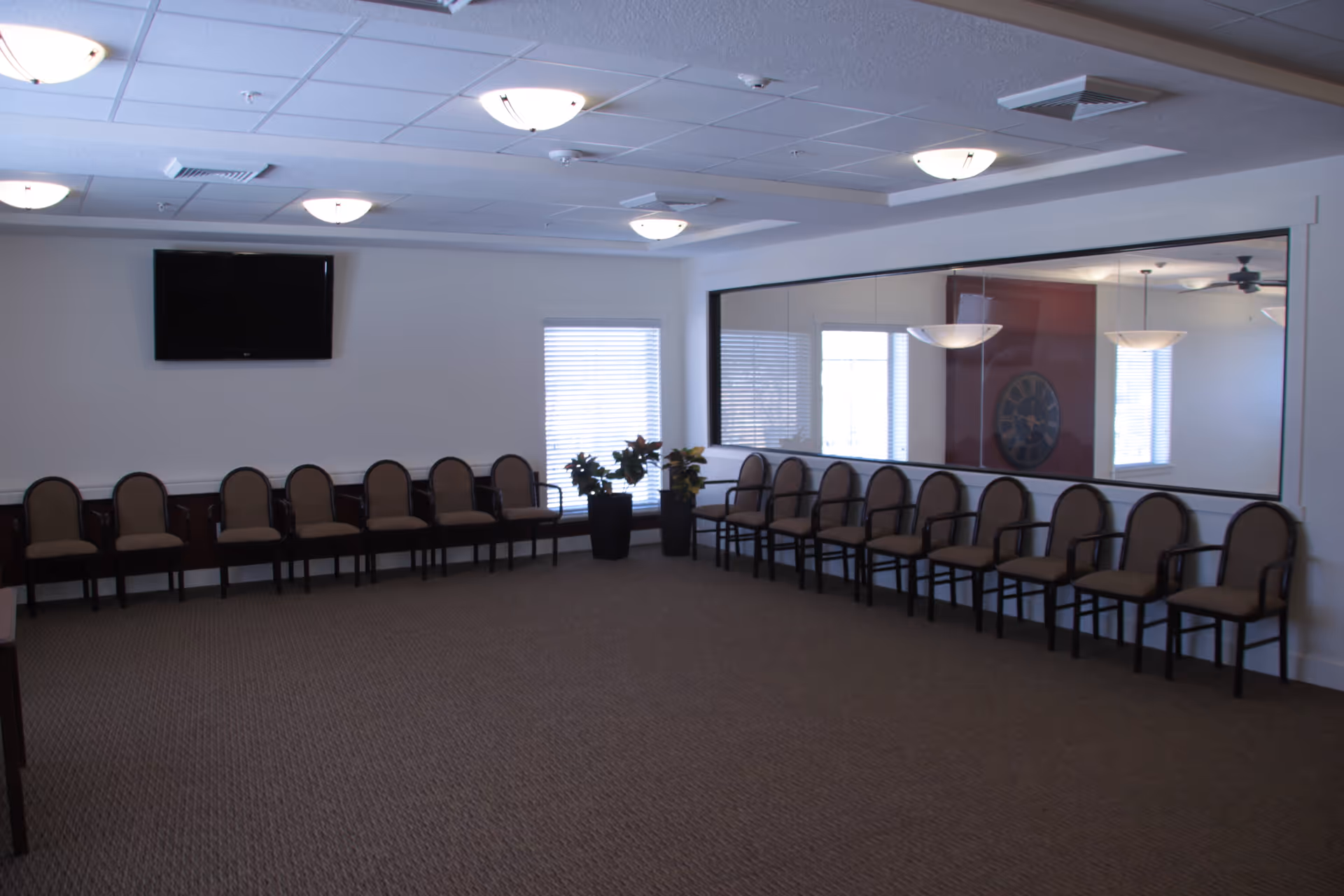 A large room with two rows of chairs lined up against the walls forming an L shape. The room has a carpeted floor, a wall-mounted flat screen TV, two windows with blinds, two potted plants, and a large interior window showing another room with hanging light fixtures and a wall clock.