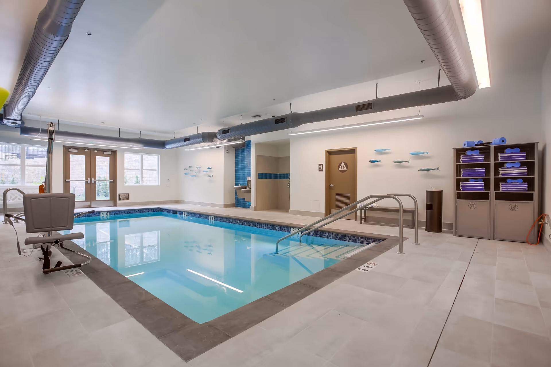 Indoor swimming pool area with clear blue water, a pool lift chair for accessibility, metal handrails, and a rack with neatly folded blue towels and pool noodles. The room has large windows and double doors letting in natural light, white walls with decorative fish wall art, and exposed ductwork on the ceiling.