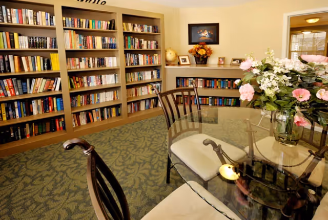 A cozy library room with built-in bookshelves filled with books along the walls. There is a glass-top table with cushioned chairs around it, and a vase of pink and white flowers on the table. The room has a patterned carpet and a framed picture on the wall.