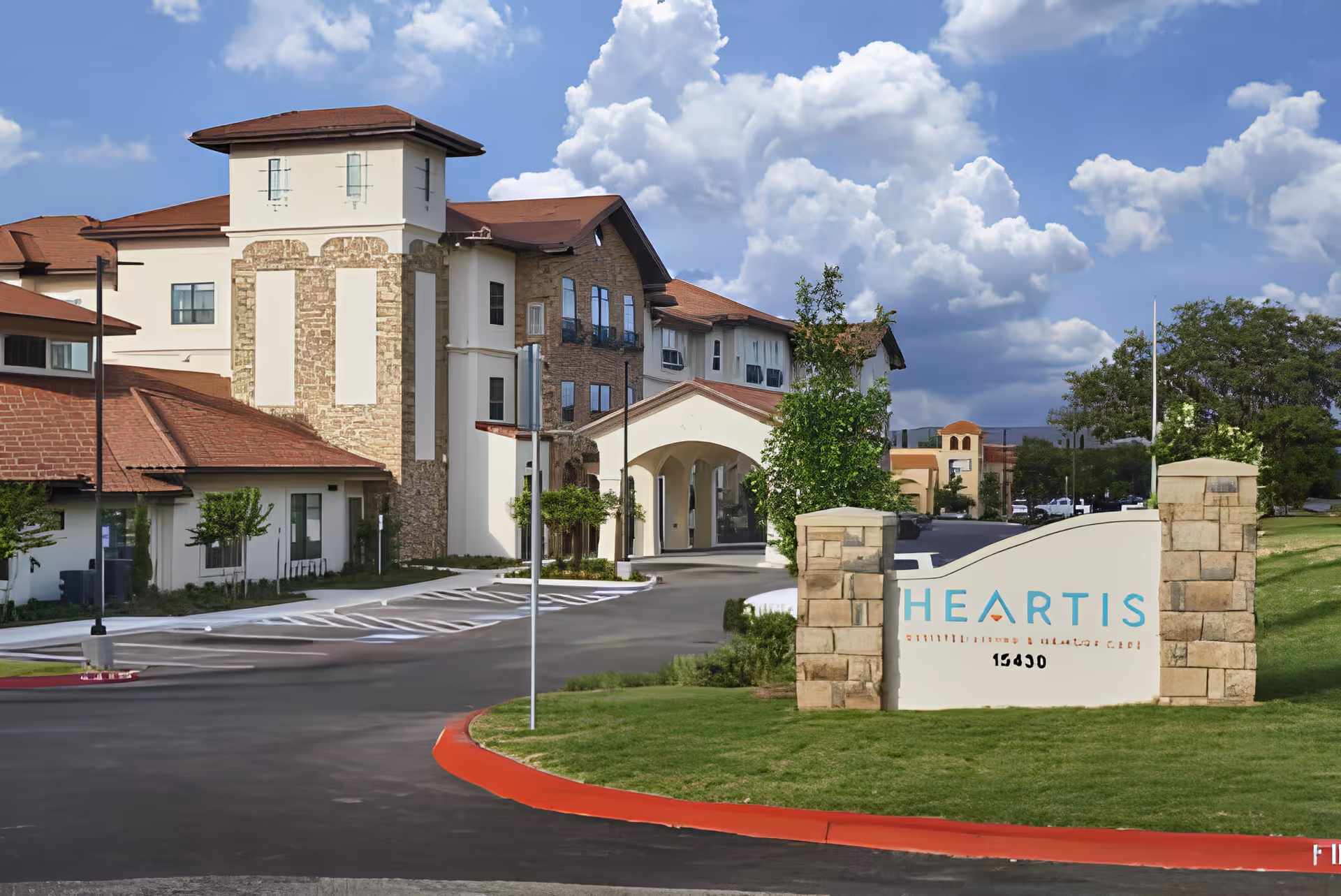 Exterior view of a senior living facility with a large building featuring a mix of stone and stucco walls, red-tiled roofs, and multiple windows. The entrance has an arched driveway with a parking area. A sign in the foreground reads 'HEARTIS' with additional smaller text and the number 15490. The sky is partly cloudy with blue patches.