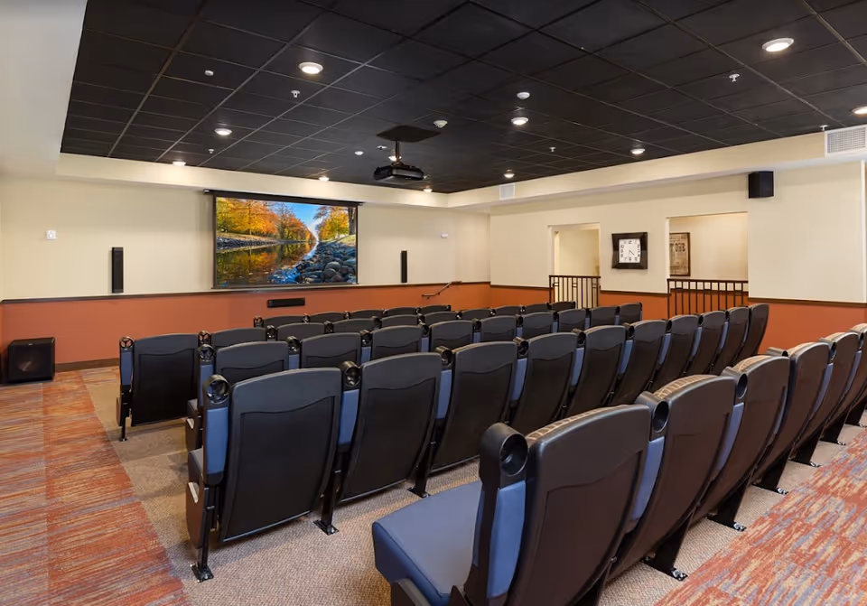 A small theater room with several rows of black and blue cushioned seats facing a large screen displaying a scenic image of a river with autumn trees. The room has a black ceiling with recessed lighting and beige walls with a reddish-brown lower section. A clock is mounted on the wall to the right.