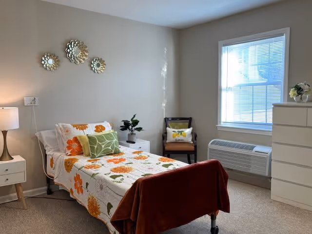 A bright and cozy bedroom with a single bed covered in a floral bedspread with orange and green flowers. There is a white nightstand with a lamp and a small plant next to the bed. On the wall above the bed are three decorative metallic flower wall hangings. A wooden chair with a cushion is placed near a window with blinds. Below the window is a white air conditioning unit. A white dresser with a small flower arrangement on top is positioned to the right.
