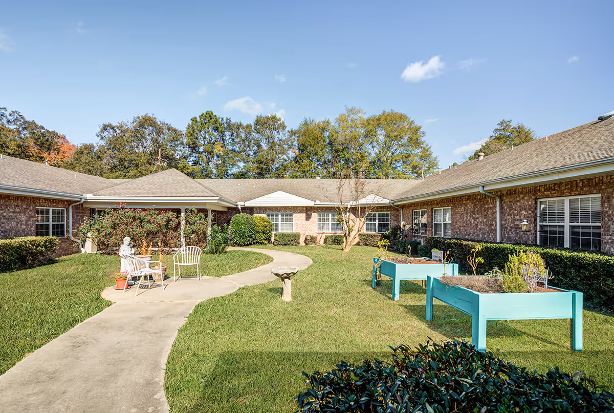 Sunny landscaped courtyard with a winding concrete path, seating, raised planters, and surrounding single-story brick buildings.