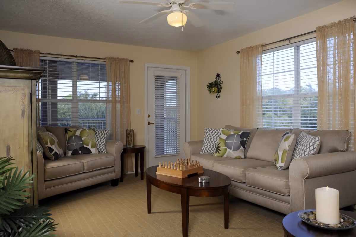 Cozy living room with two beige sofas adorned with patterned throw pillows, a wooden coffee table with a chess set, windows with blinds and curtains, and a ceiling fan.
