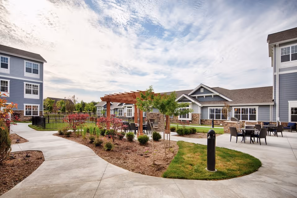 Courtyard of a senior living facility with paved walkways, a wooden pergola, patio seating, landscaping, and surrounding buildings under a partly cloudy sky.