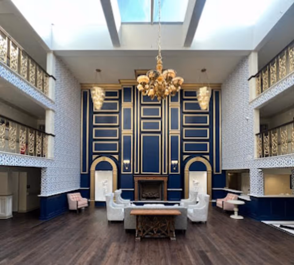 A spacious and elegant living room area with high ceilings and a large skylight. The room features a dark wood floor, a central seating arrangement with white and pink upholstered chairs around a wooden coffee table, and a decorative fireplace on a navy blue and gold accent wall. Ornate gold railings line the balconies on both sides of the room, and a large chandelier hangs from the ceiling.