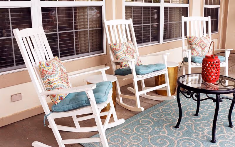 A cozy outdoor seating area with three white wooden rocking chairs, each with a blue cushion and a colorful patterned pillow. There is a small round glass-top table with a red decorative lantern on it, and a blue rug with a swirling pattern on the floor. The area is adjacent to a building with large windows and beige walls.