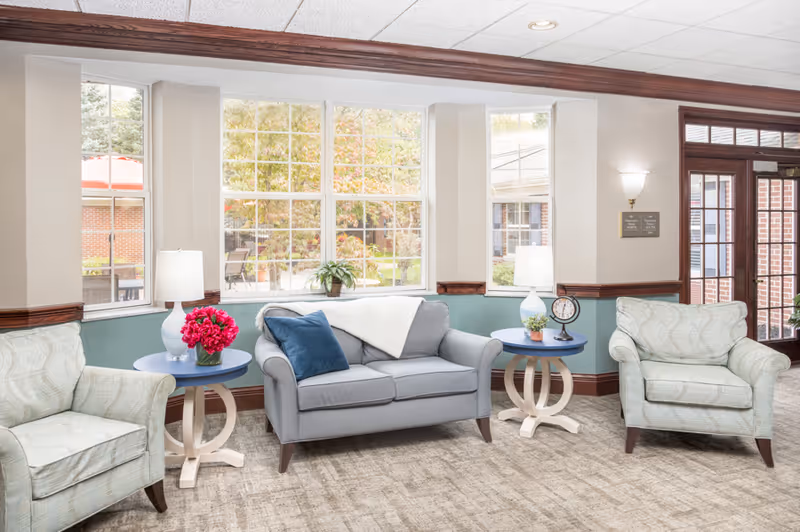A bright and cozy sitting area in a senior living facility with a large window showing outdoor greenery. The room features a gray loveseat with a blue pillow and a white throw, flanked by two light green armchairs. Two round side tables hold white lamps, a clock, a small plant, and a vase with pink flowers. The walls are painted in a soft beige and teal color with wood trim, and there is a glass door on the right side.
