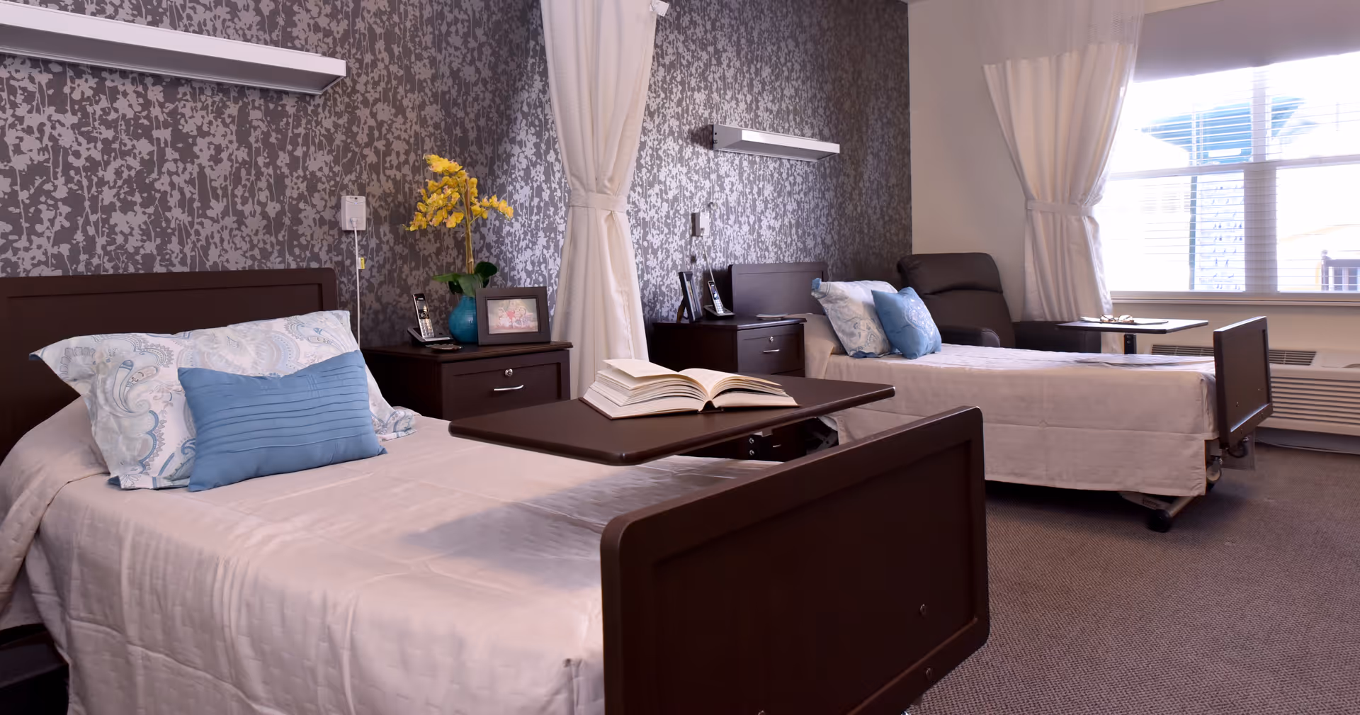 A well-lit room with two single beds, each with white bedding and decorative pillows. Between the beds are two dark wooden nightstands, one with a vase of yellow flowers and a framed photo. A book is open on a movable overbed table on the bed in the foreground. The walls have a patterned wallpaper, and large windows with white curtains allow natural light to fill the room. There is also a dark armchair near the window.