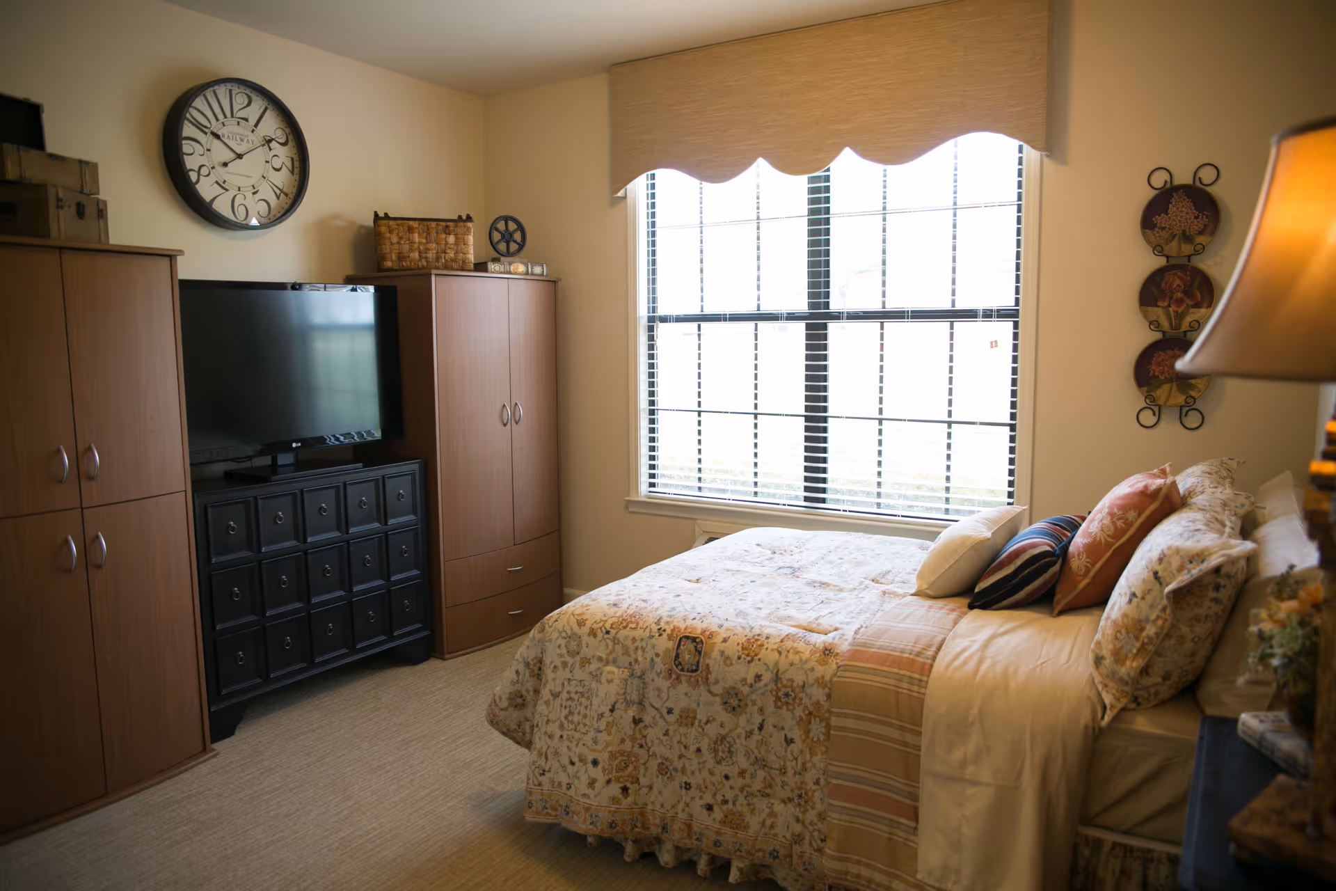A cozy bedroom with a made bed and decorative pillows, a large window with blinds, and cabinetry with a TV and clock.