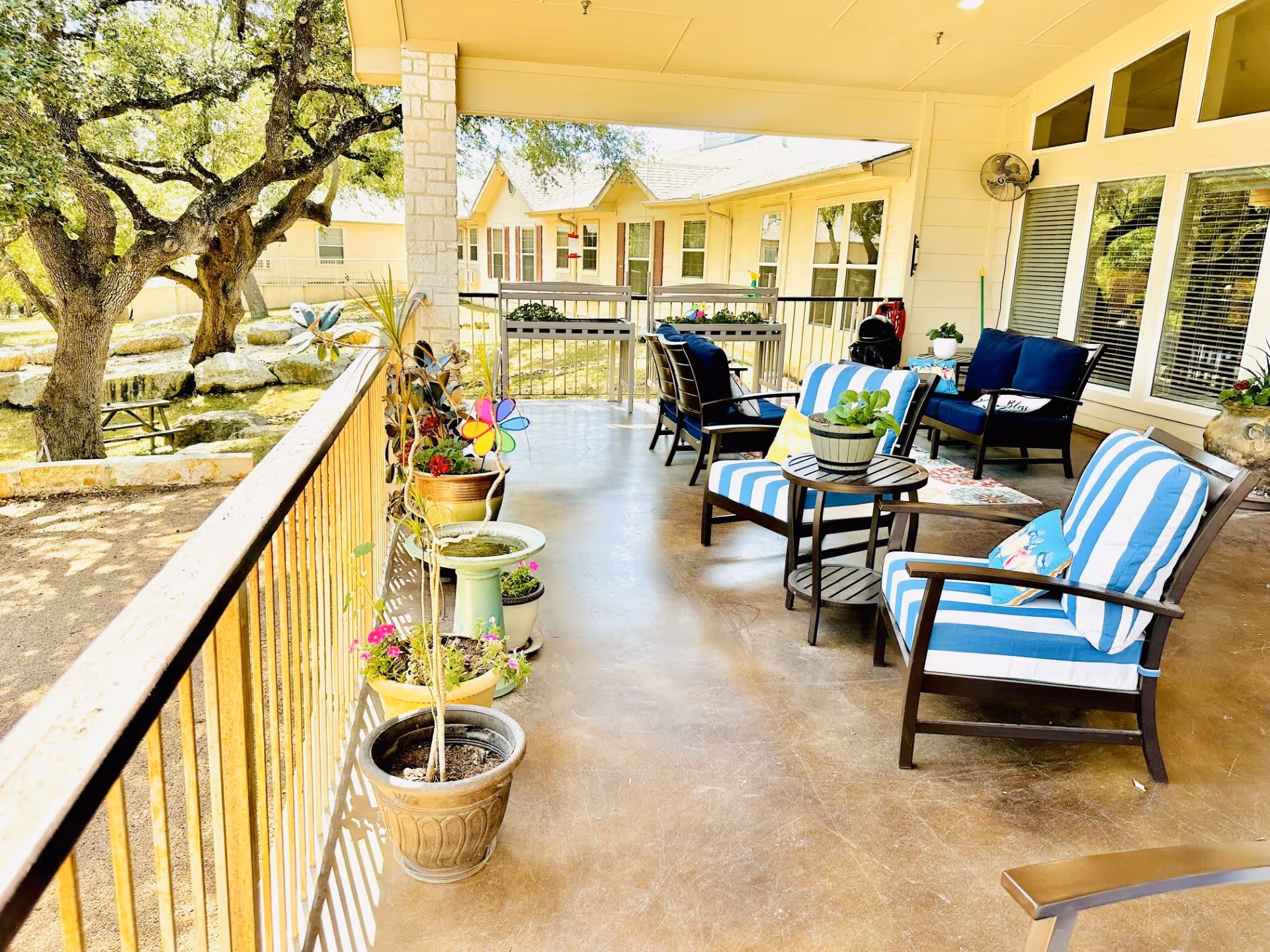 A covered outdoor patio area at Heritage Place of Boerne with several cushioned chairs featuring blue and white striped cushions, small tables, and various potted plants along the railing. Large trees and a picnic table are visible outside the patio area.