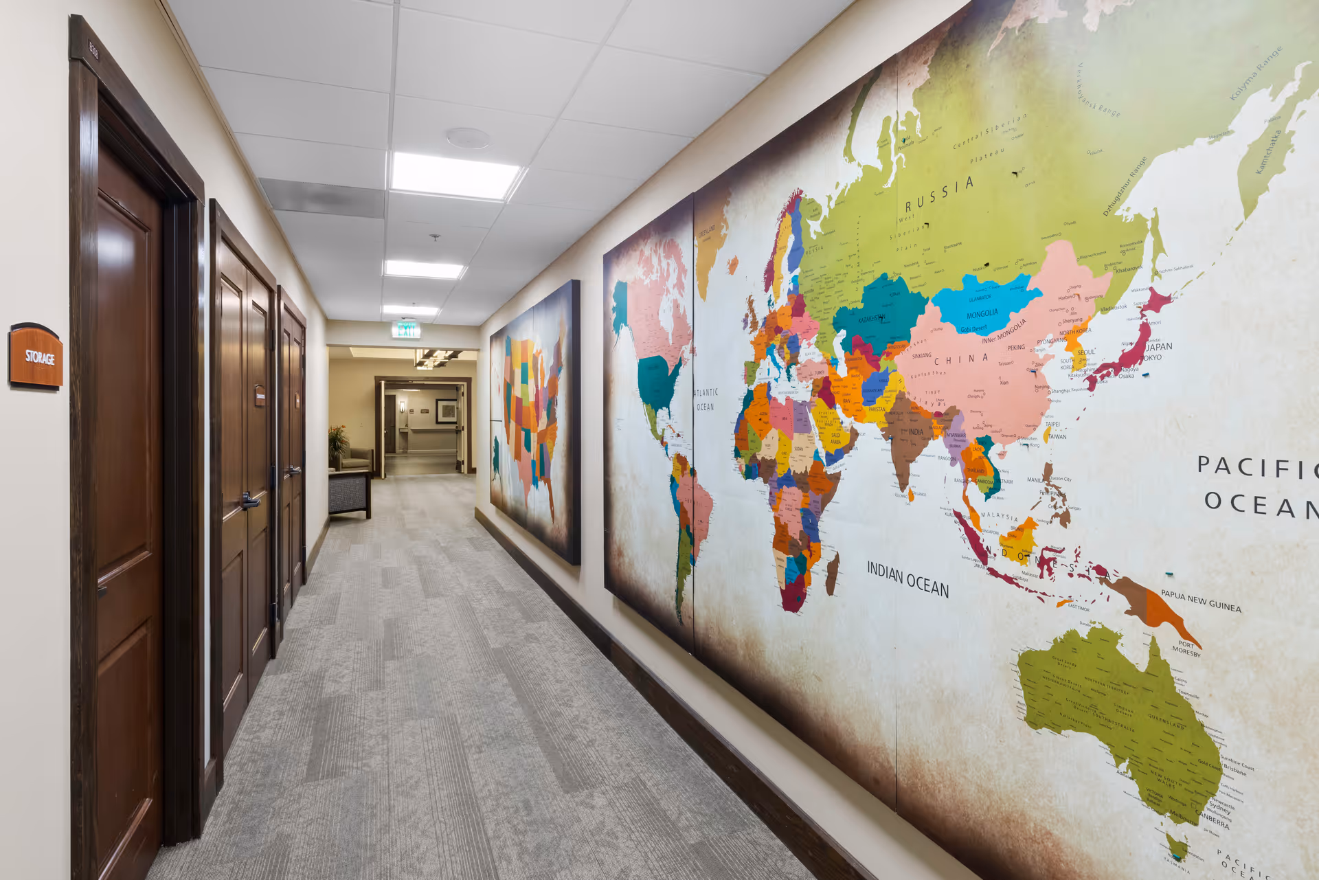 A well-lit hallway in a senior living facility with carpeted floors and wooden doors on the left side. The right wall features large colorful maps of the world and the United States. A sign labeled 'STORAGE' is visible on one of the doors.