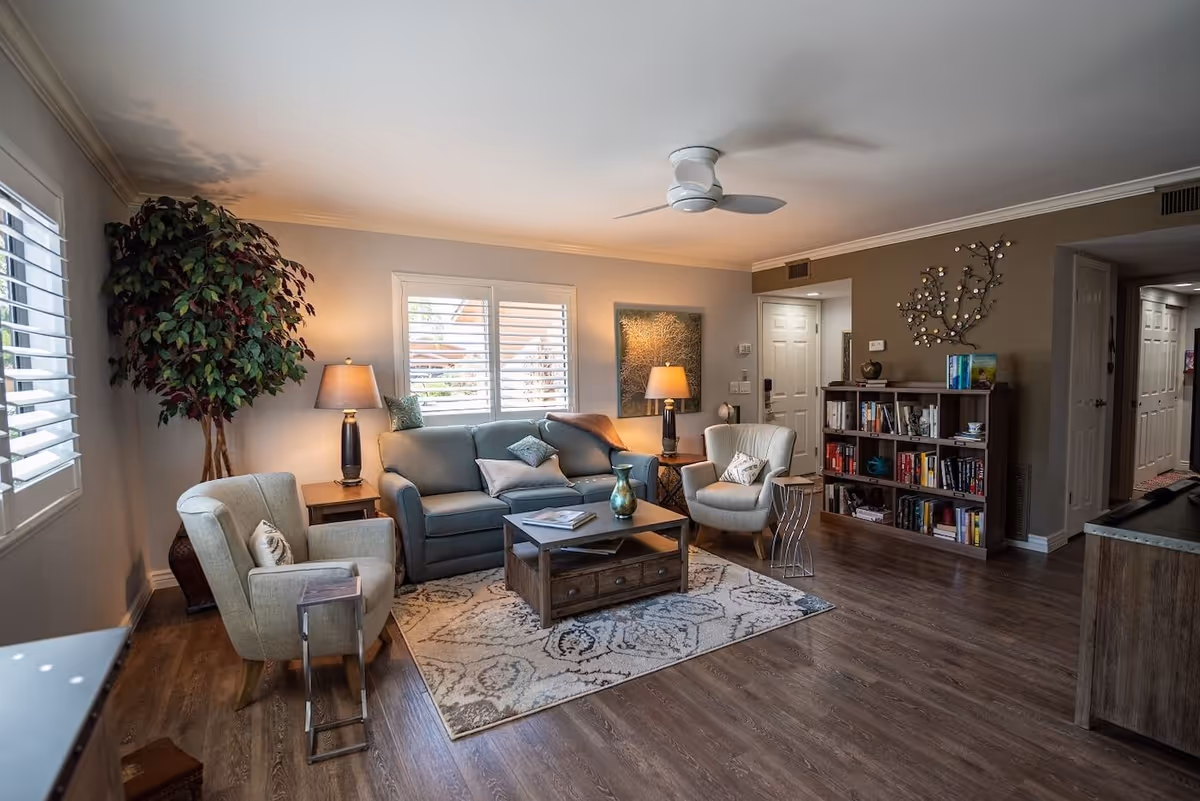A cozy living room with a blue sofa, two beige armchairs, a wooden coffee table on a patterned rug, two table lamps, a large potted plant, a bookshelf filled with books, and decorative wall art. The room has wooden flooring and a ceiling fan.