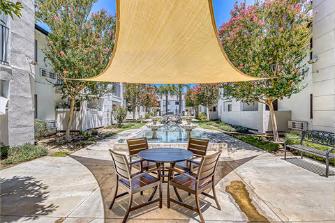 Outdoor courtyard area at Parkside Senior Apartments featuring a round table with four chairs under a large beige shade sail. The courtyard is lined with flowering trees and white apartment buildings on both sides, with a water feature in the center background.