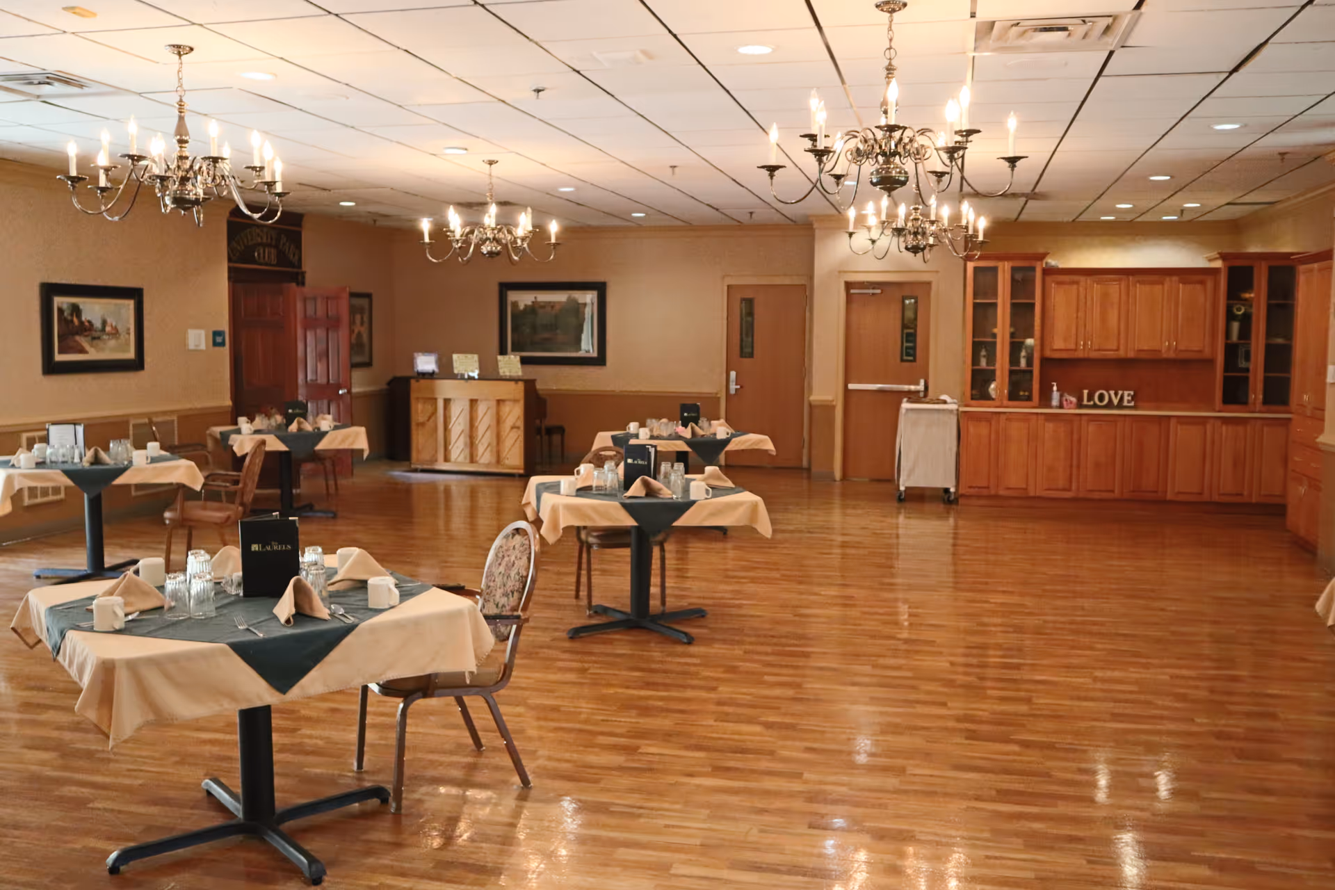 A spacious dining room with wooden floors and beige walls, featuring several tables set with beige tablecloths and green overlays, napkins, cups, and glasses. The room is illuminated by multiple chandeliers hanging from a white ceiling. There are wooden cabinets along one wall with decorative items and the word 'LOVE' displayed. Paintings hang on the walls, and a piano is visible in the background near a wooden door.