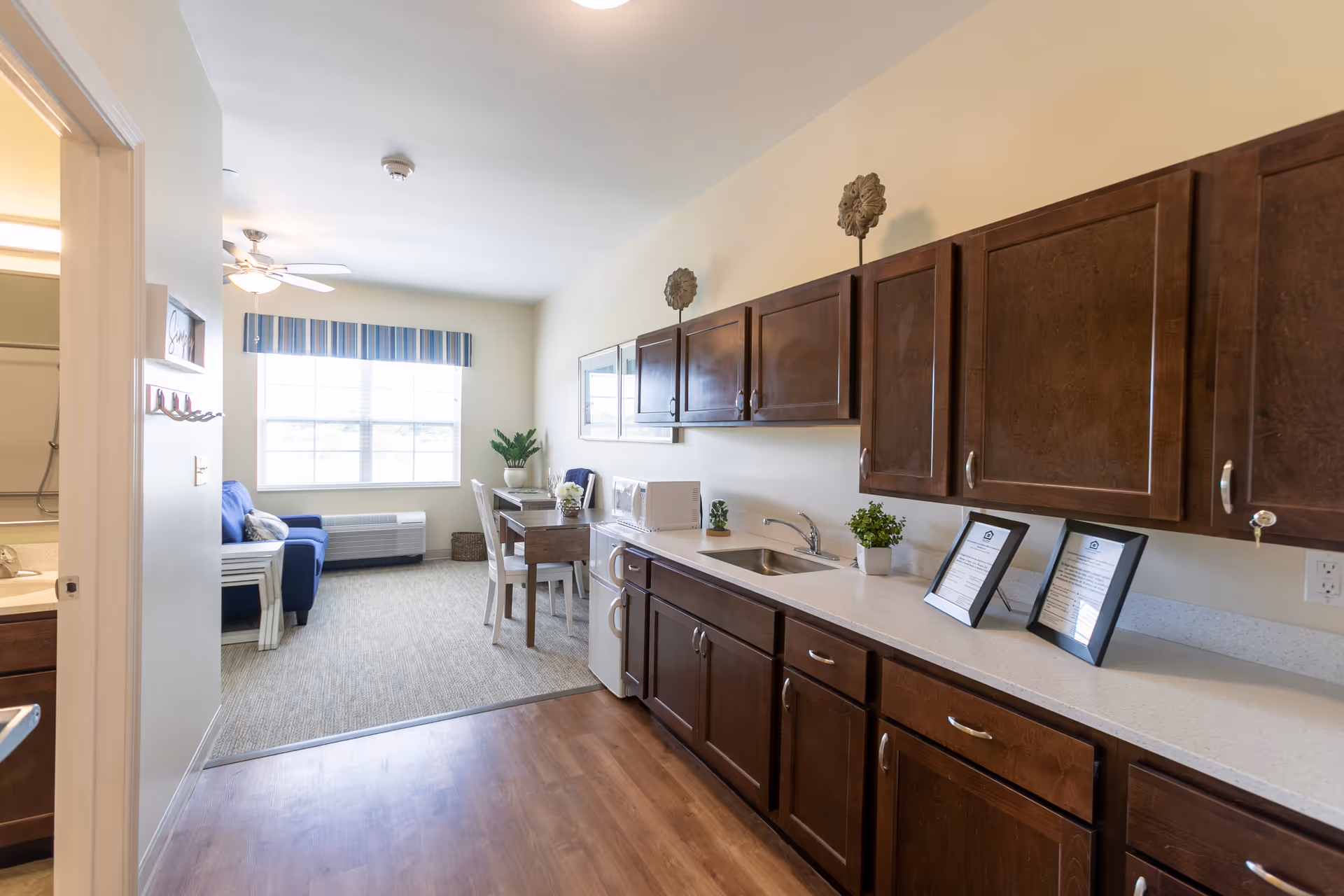 Small apartment kitchenette with dark wood cabinets and a sink opening into a sitting area with a dining table, window, and sofa.
