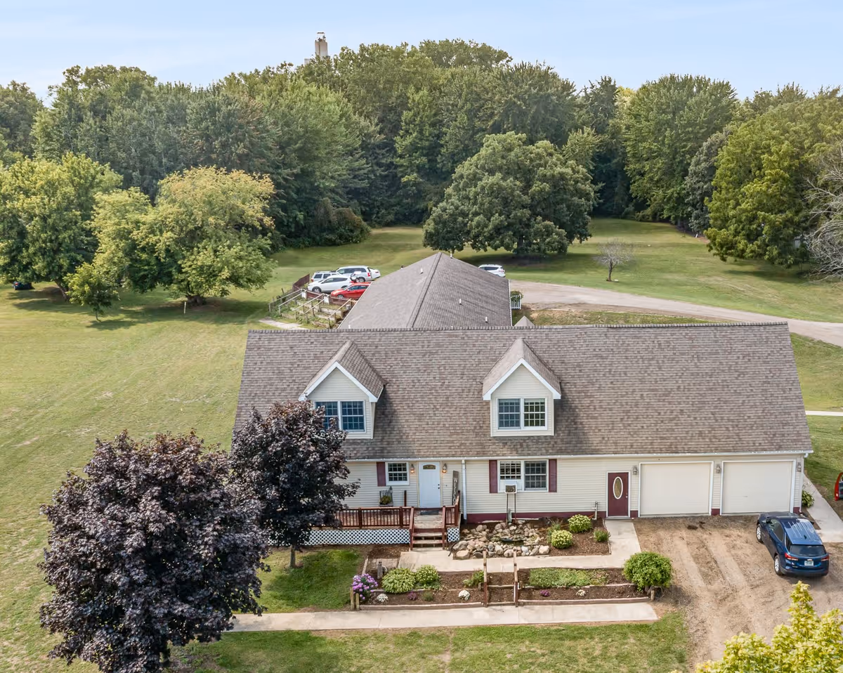 Aerial view of a single-story assisted living facility building with a gable roof, surrounded by green lawns and trees. There are several cars parked near the building, and a paved walkway leads to the entrance. The area is spacious with a backdrop of dense trees under a clear sky.