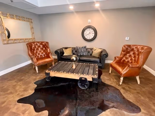 A cozy sitting area with a black leather sofa adorned with cushions, two brown leather tufted armchairs, a rustic wooden coffee table on wheels, and a dark animal hide rug on the floor. A large round wall clock is mounted on the beige wall above the sofa, and a rectangular mirror with a decorative frame hangs on the adjacent wall.