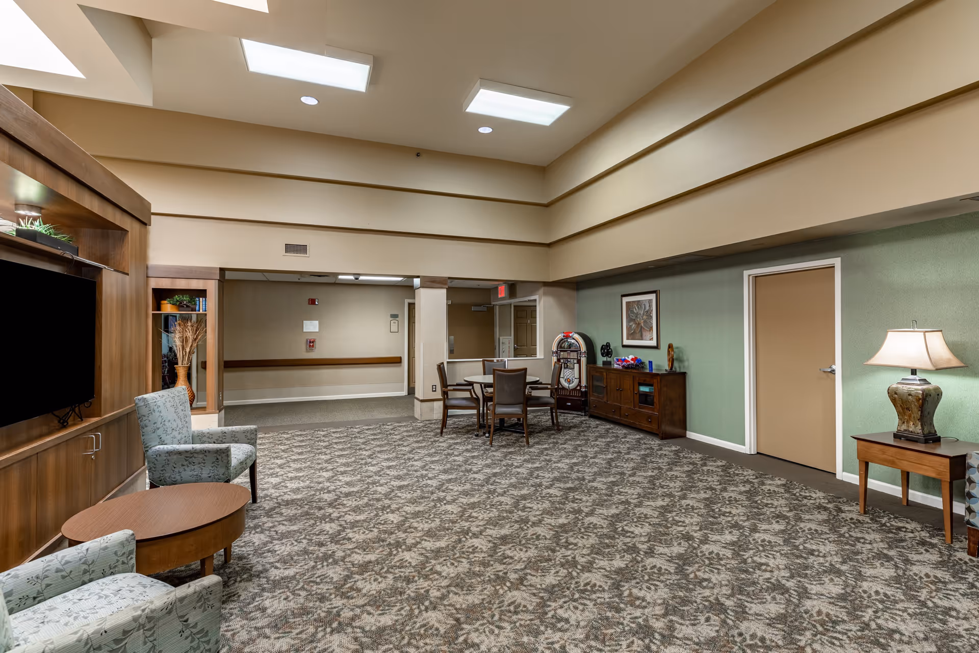 A spacious senior living community common area with patterned carpet, two upholstered armchairs, a wooden coffee table, a large flat-screen TV mounted on a wooden entertainment unit, a round table with four chairs, a vintage jukebox, a wooden sideboard with decorative items, a framed artwork on a green wall, and a table lamp on a small wooden side table.