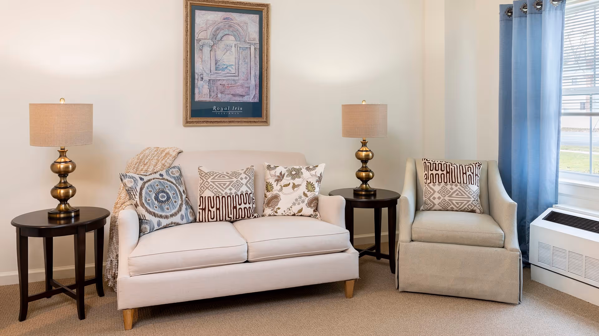 A cozy living room area with a beige sofa adorned with three decorative pillows and a throw blanket. Next to the sofa is a round wooden side table with a brass lamp. To the right of the sofa is a beige armchair with a patterned pillow, another round wooden side table with a matching brass lamp, and a window with blue curtains letting in natural light. A framed artwork hangs on the wall above the sofa.