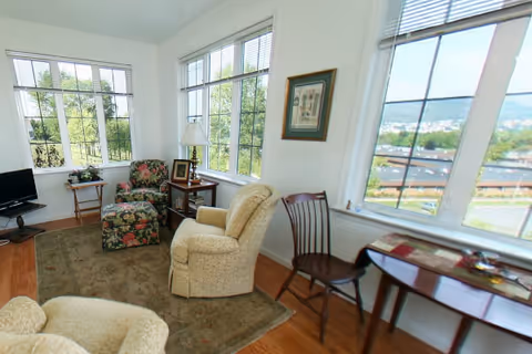 A bright living room with large windows showing an outdoor view. The room features two beige armchairs, a floral patterned armchair with matching ottoman, a small wooden side table with a lamp and picture frame, a wooden chair, a small table with magazines, and a television on a stand. The floor is hardwood with a large area rug.
