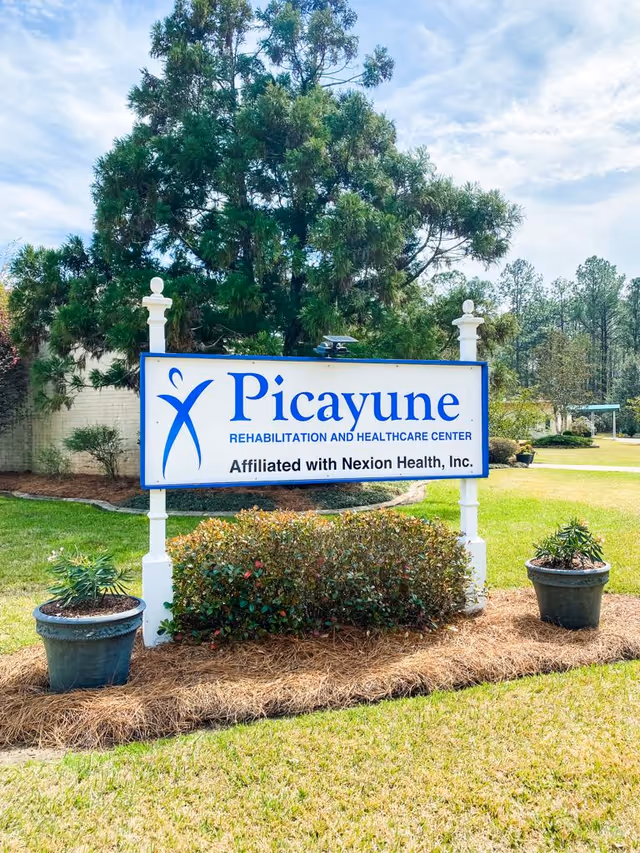 Outdoor view of a sign for Picayune Rehabilitation and Healthcare Center, affiliated with Nexion Health, Inc., surrounded by greenery, potted plants, and trees under a partly cloudy sky.
