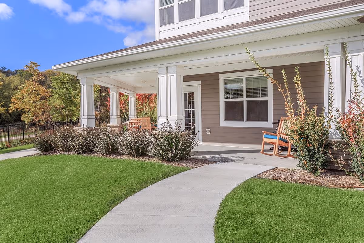 Covered porch area of a building with white columns, beige siding, and a concrete walkway curving through a well-maintained green lawn. There are wooden chairs on the porch and shrubs planted along the edge. Trees with autumn foliage are visible in the background under a partly cloudy blue sky.