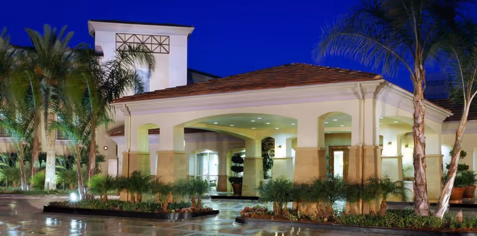Exterior view of The Village at Sherman Oaks building entrance at dusk, featuring a covered driveway with columns, palm trees, and well-maintained landscaping under a deep blue sky.