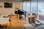 Dining area with tables set for meals, featuring white tablecloths, plates, and silverware. A black grand piano is positioned near large windows with blinds, allowing natural light to fill the room. Wooden chairs with striped cushions surround the tables, and the floor is made of light-colored wood.