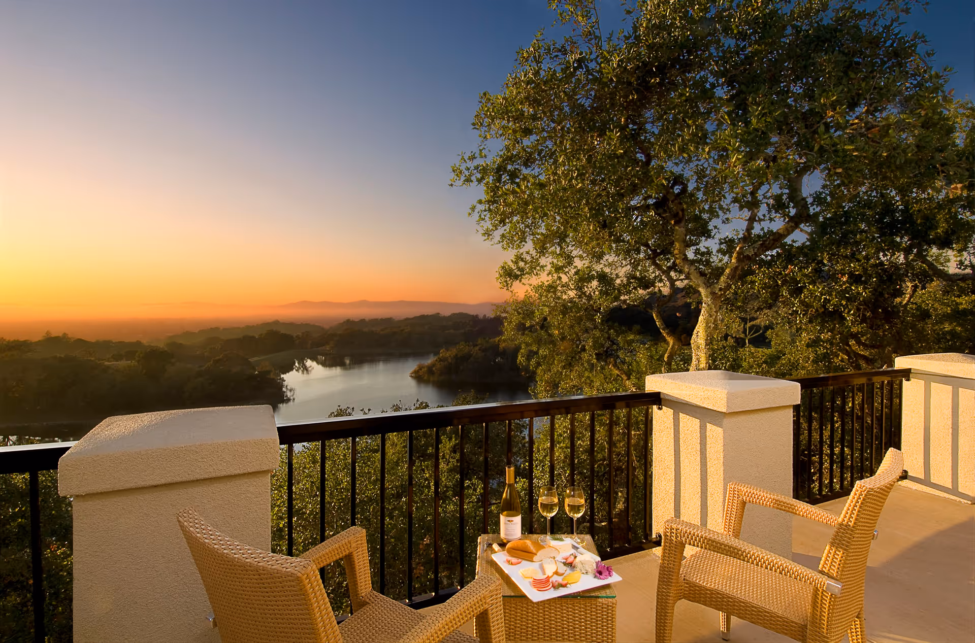 Balcony with two wicker chairs and a small table holding wine and snacks overlooking a lake and trees at sunset.