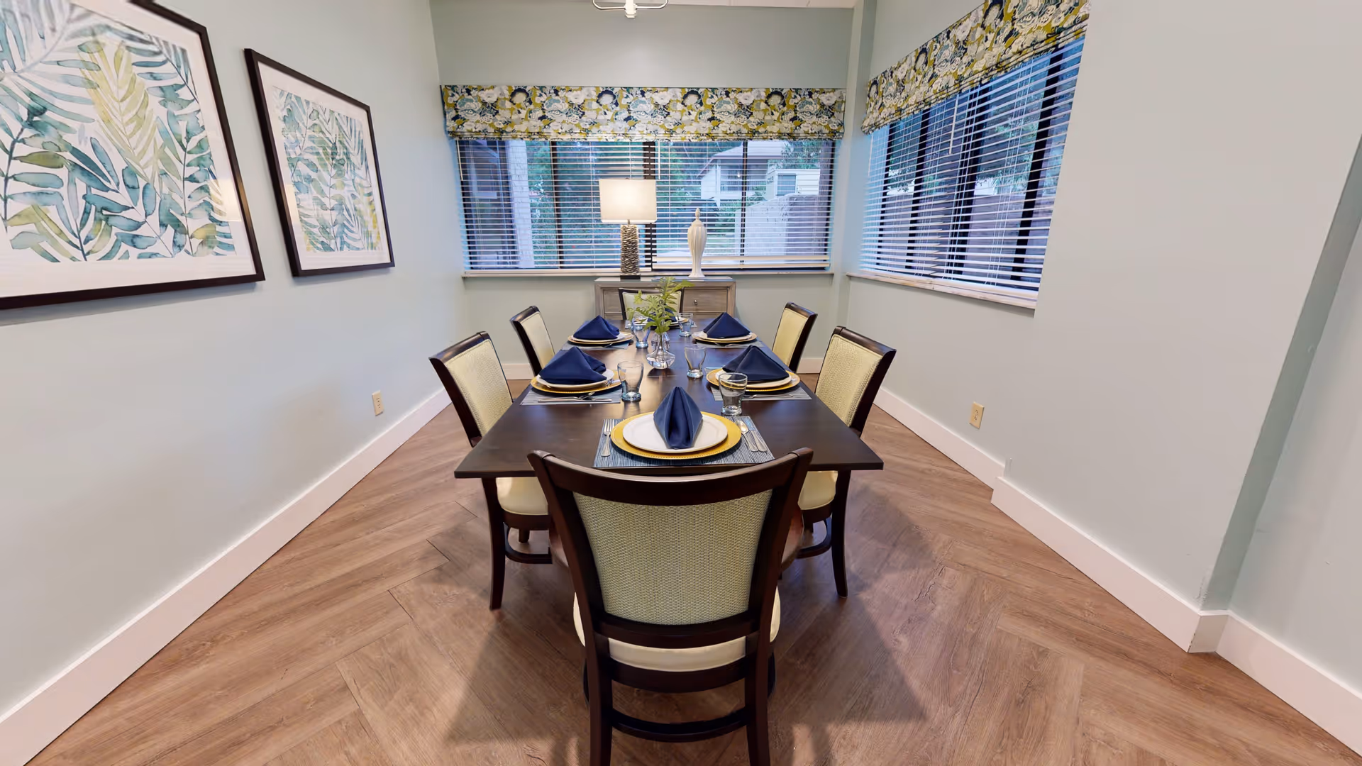 A dining room with a rectangular dark wood table set for six people with blue napkins, plates, and glasses. The room has light-colored walls, two framed botanical prints on one wall, and two windows with floral valances letting in natural light. A lamp and decorative vase are placed on a sideboard under the windows.