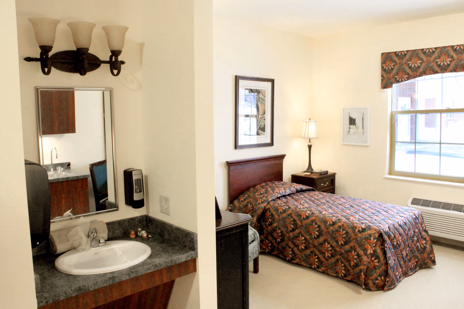 A senior living facility room featuring a single bed with a patterned bedspread, a nightstand with a lamp, framed artwork on the walls, a window with a valance, and a small sink area with a mirror and soap dispenser.