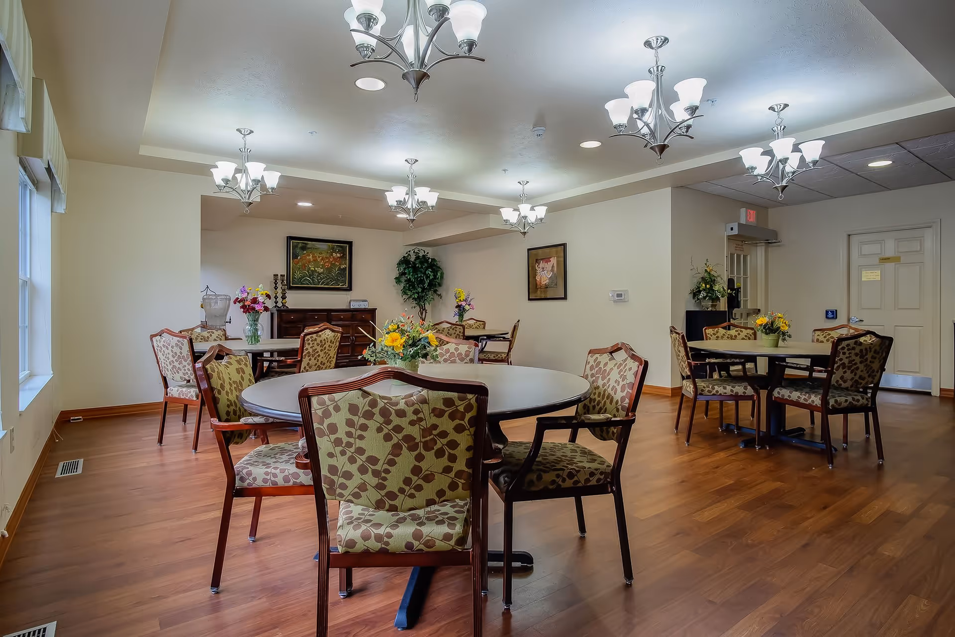 A well-lit dining room with several round tables surrounded by cushioned chairs featuring a leaf pattern. The room has wooden flooring, multiple ceiling light fixtures, framed artwork on the walls, and floral centerpieces on the tables. There is a window on the left side letting in natural light.