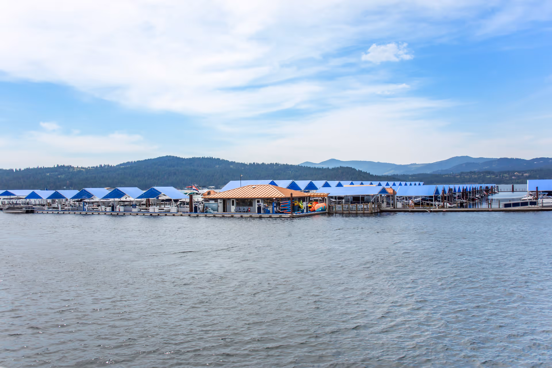 A marina with multiple covered boat slips featuring blue roofs, situated on a body of water with forested hills and mountains in the background under a partly cloudy sky.
