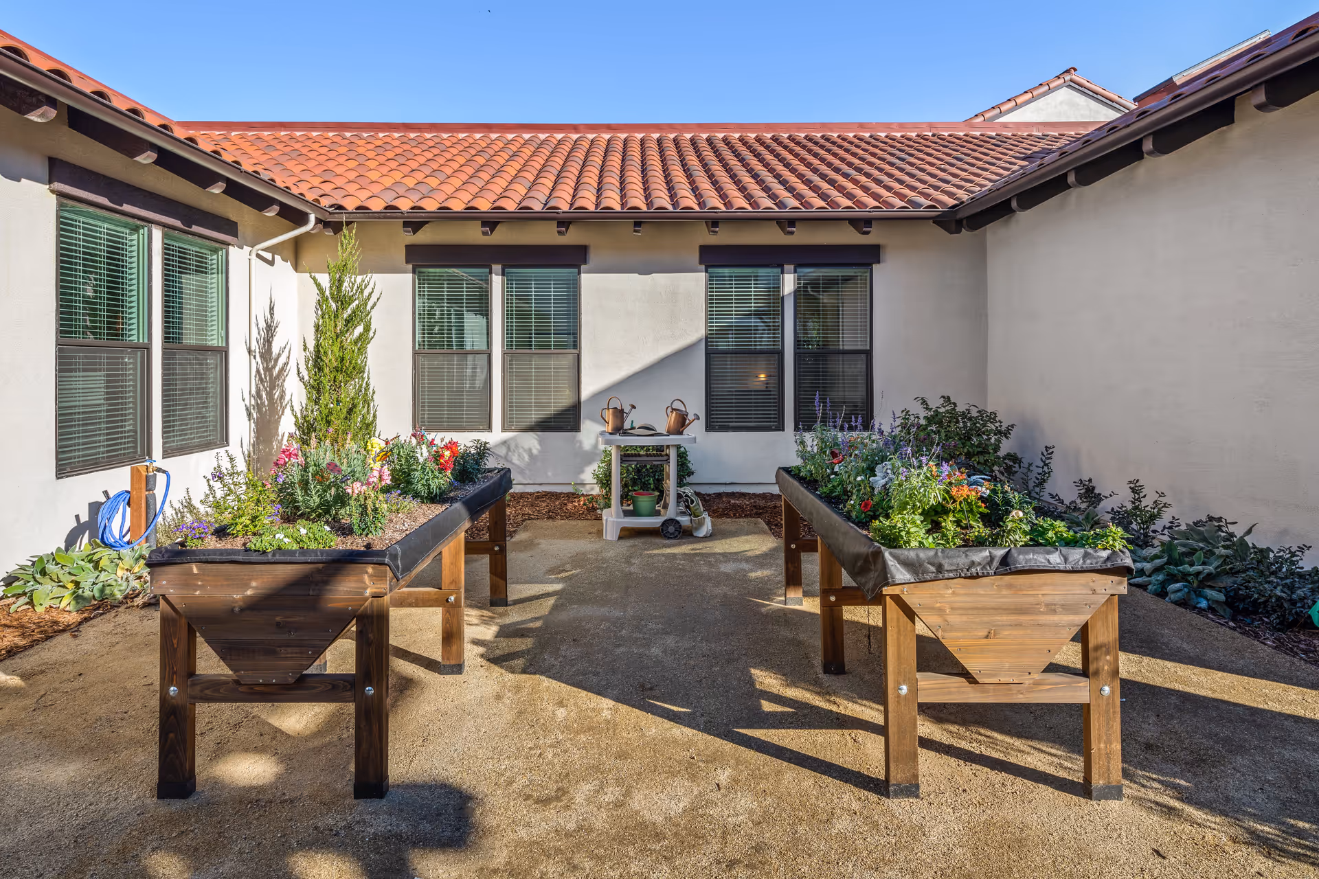 Outdoor courtyard area with two raised wooden garden beds filled with various plants and flowers. The courtyard is surrounded by beige walls with windows and a red tile roof. A small cart with watering cans and gardening supplies is positioned against the far wall.
