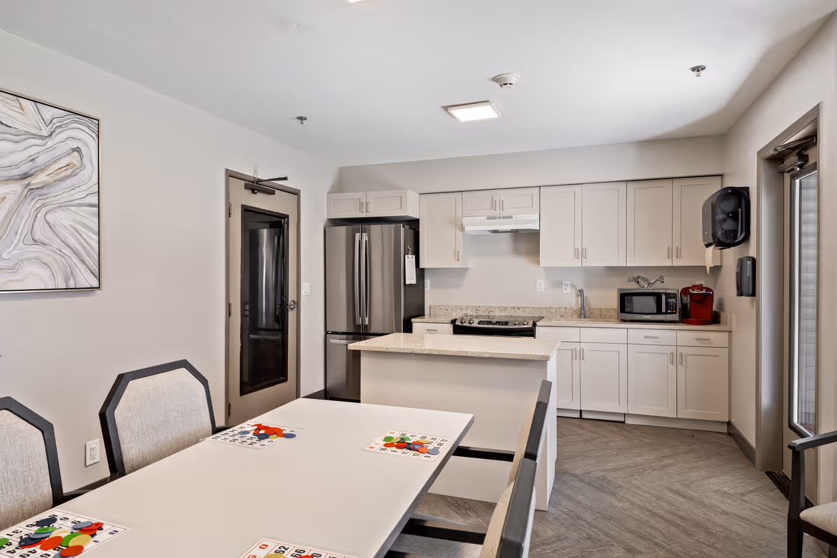 A modern kitchen and dining area in a senior living community. The kitchen features white cabinets, a stainless steel refrigerator, a stove with a range hood, a microwave, and a red coffee maker. There is a small island with a countertop in the center. The dining table in the foreground has chairs around it and bingo cards with colorful markers placed on the table. The walls are light-colored, and there is abstract artwork on one wall. A door with a glass panel is visible near the refrigerator, and another door leads outside.