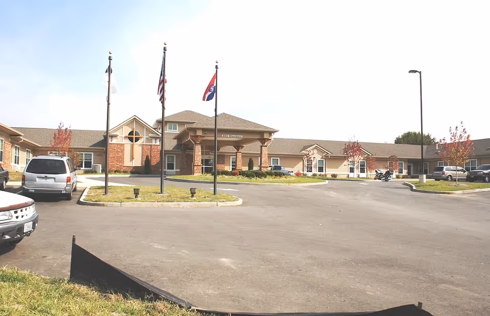Front exterior view of Christian Care Home building with a driveway, three flagpoles with flags, parked cars, and small trees with red leaves.