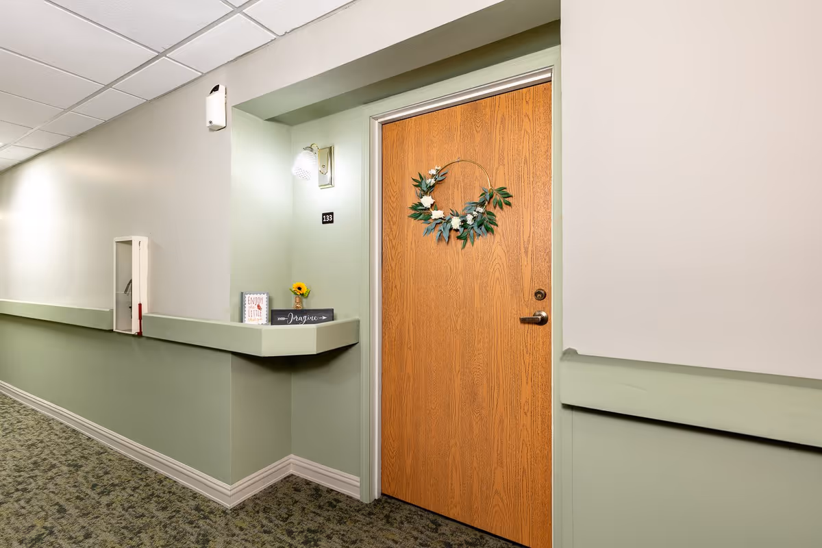 A hallway in a senior living facility with light green and beige walls, carpeted floor, and a wooden door decorated with a floral wreath. The door is labeled with the number 133. A small shelf next to the door holds a small sunflower in a vase and two decorative signs, one reading 'Enjoy the little things' and the other 'Imagine'. A wall-mounted light fixture is above the door.