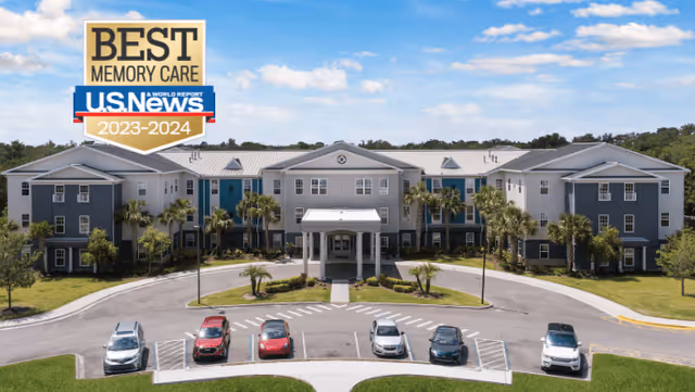 Front exterior view of a large, three-story senior living facility building with a covered entrance, palm trees, and a parking area with several cars. The sky is blue with some clouds. A badge in the top left corner reads 'Best Memory Care U.S. News 2023-2024'.