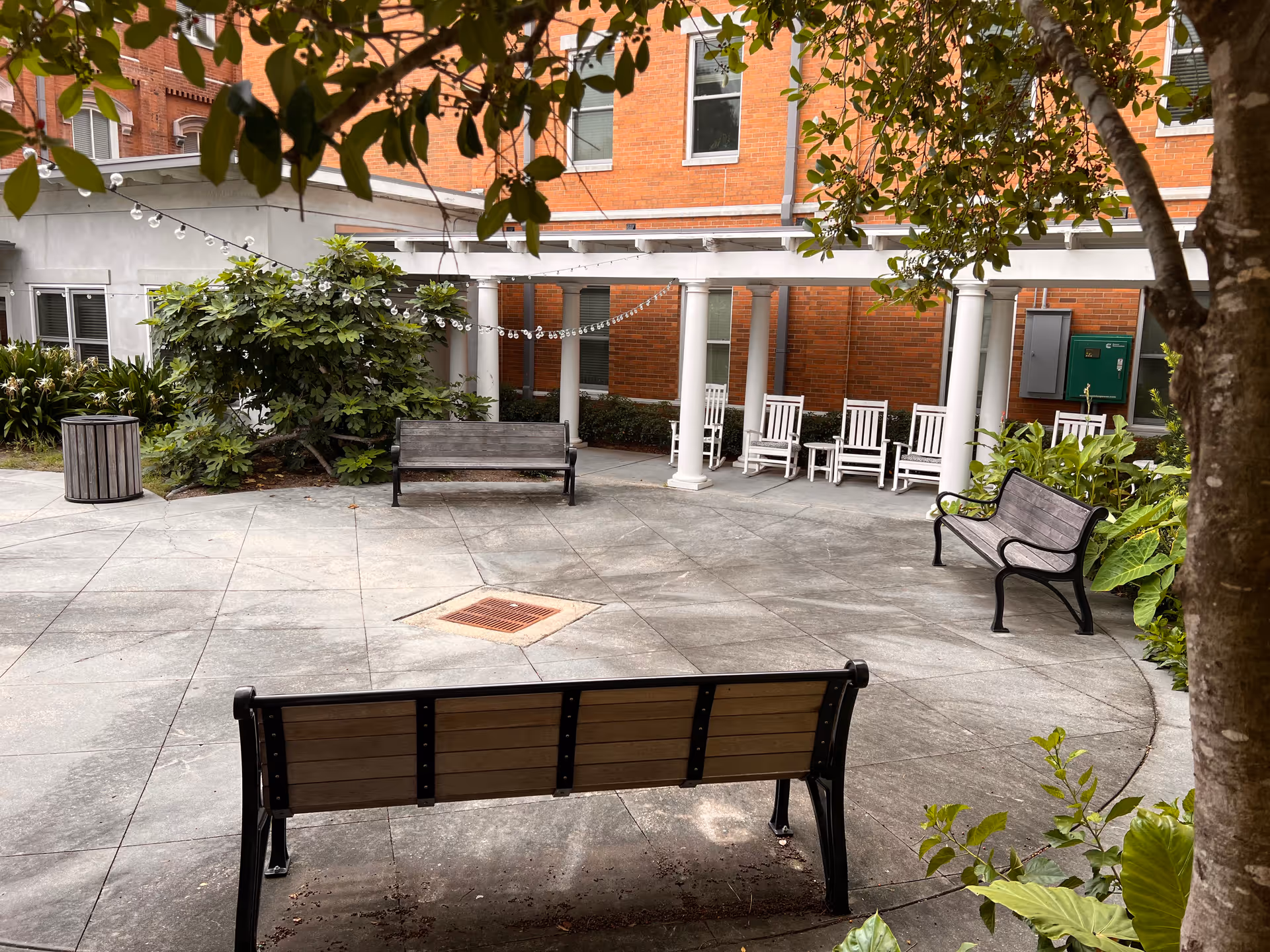 Outdoor courtyard area with several benches and white rocking chairs under a pergola. The space is surrounded by greenery and trees, with a brick building in the background. String lights are hung across the pergola.