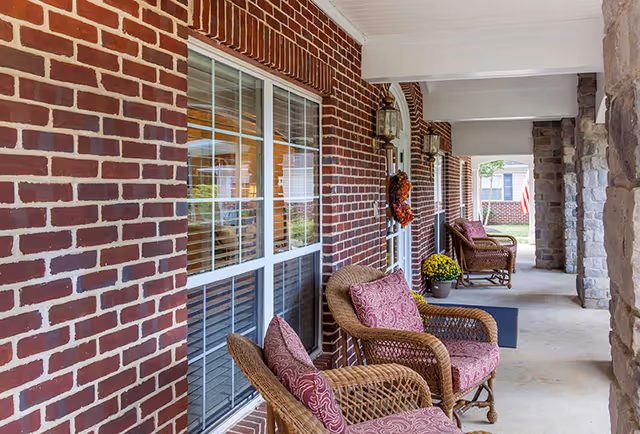 Covered brick front porch with wicker chairs topped with patterned cushions, potted flowers, and stone columns.