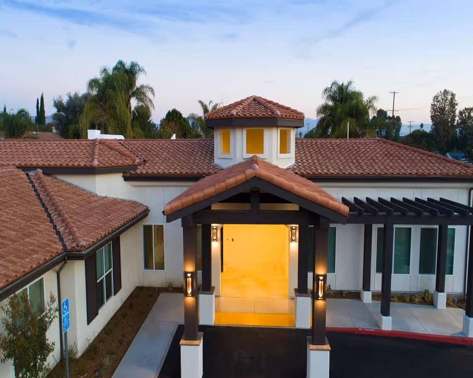 Exterior view of The Preserve at Woodland Hills Assisted Living & Memory Care building entrance during dusk, featuring a covered entryway with warm lighting, beige walls, and a red tile roof surrounded by palm trees.